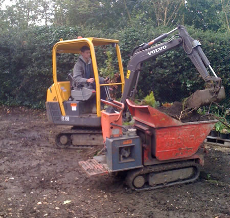 A mini digger loading soil into a tracked dumper on a cleared garden plot.